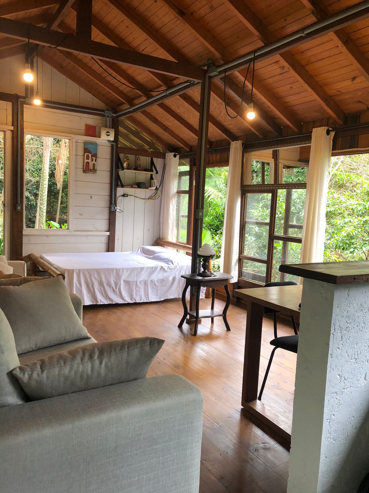 A cozy interior features a spacious bed covered with a white blanket, surrounded by large windows that offer views of the lush greenery outside. A small table and two chairs are positioned nearby, along with a comfortable sofa and decorative accents above the bed.