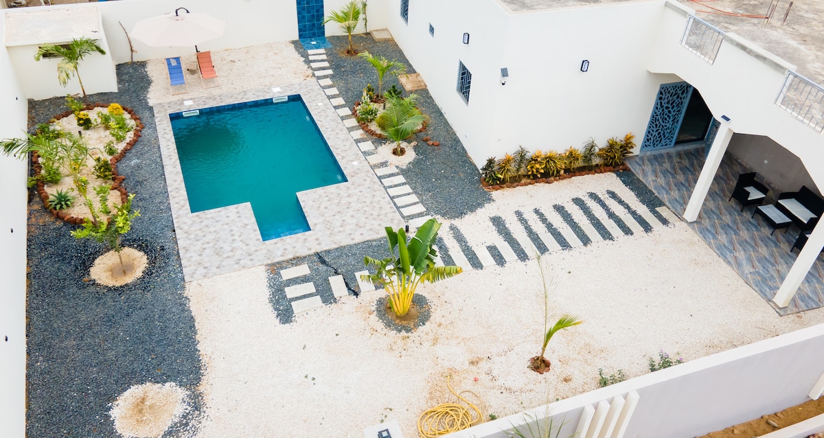An aerial view of the outdoor area features a rectangular pool surrounded by a stone patio. Pathways of large stones are visible, leading through the landscaped garden with various plants and a shaded seating area under an umbrella.
