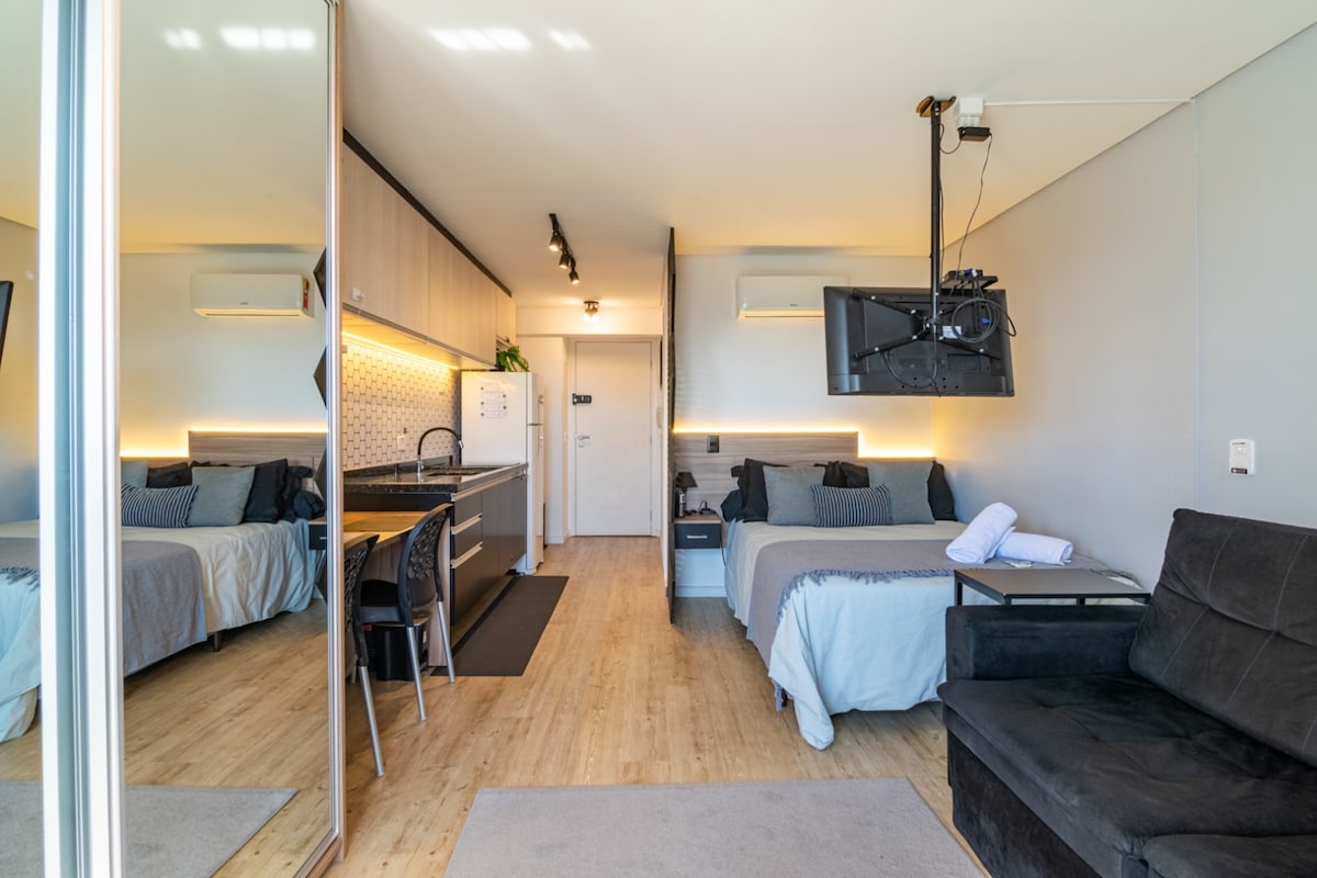 A compact studio features a well-organized kitchen with modern appliances, visible from the cozy sleeping area. A dark sofa is positioned next to a bed adorned with neutral-toned linens. Natural light floods through the large mirror, reflecting the open layout of the space.