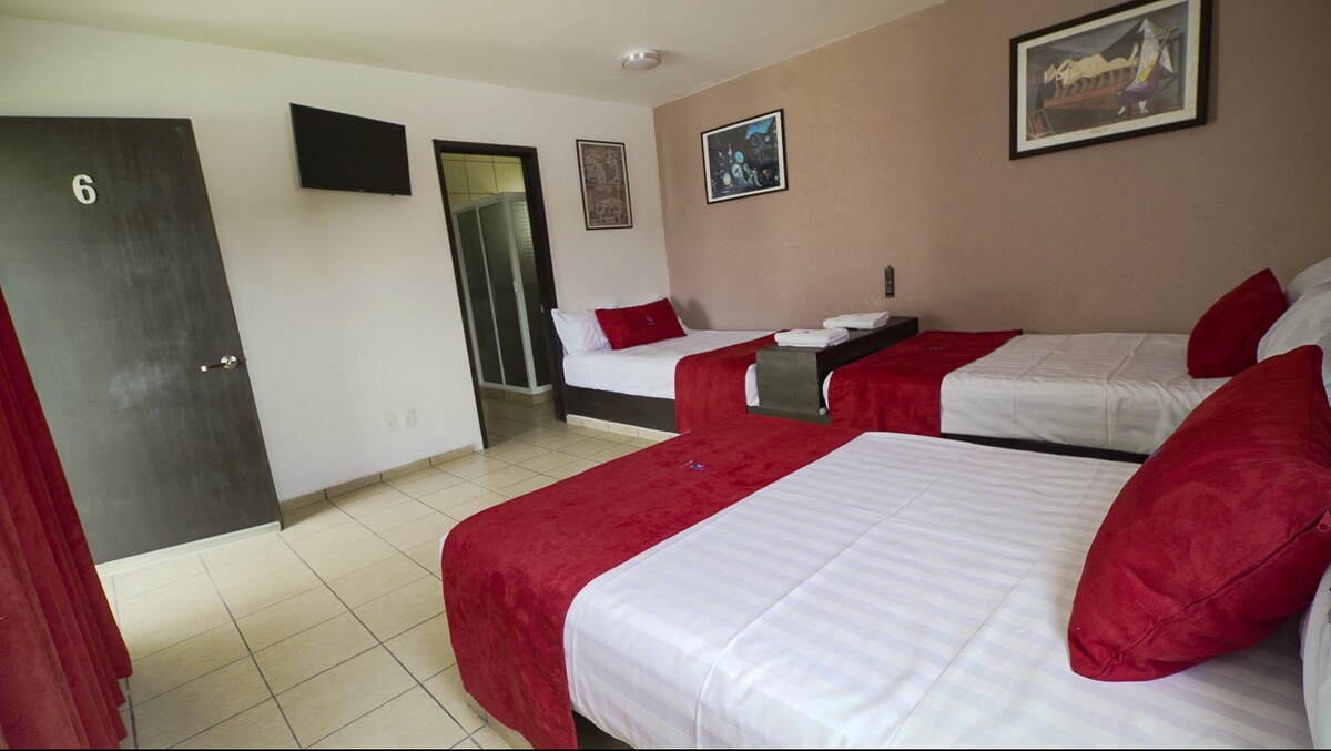 The room features three spacious queen-size beds with red and white linens. A flat-screen TV is mounted on the wall, and a bathroom door is visible in the background. Artwork is displayed above the beds, enhancing the room's decor.