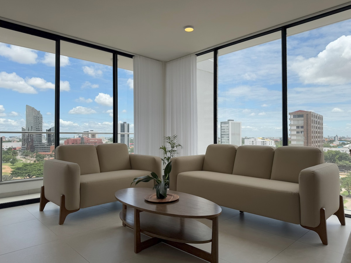 A modern living area features two light-colored sofas positioned around a round wooden coffee table. Floor-to-ceiling windows provide ample natural light and showcase an expansive view of the city skyline and clouds above.