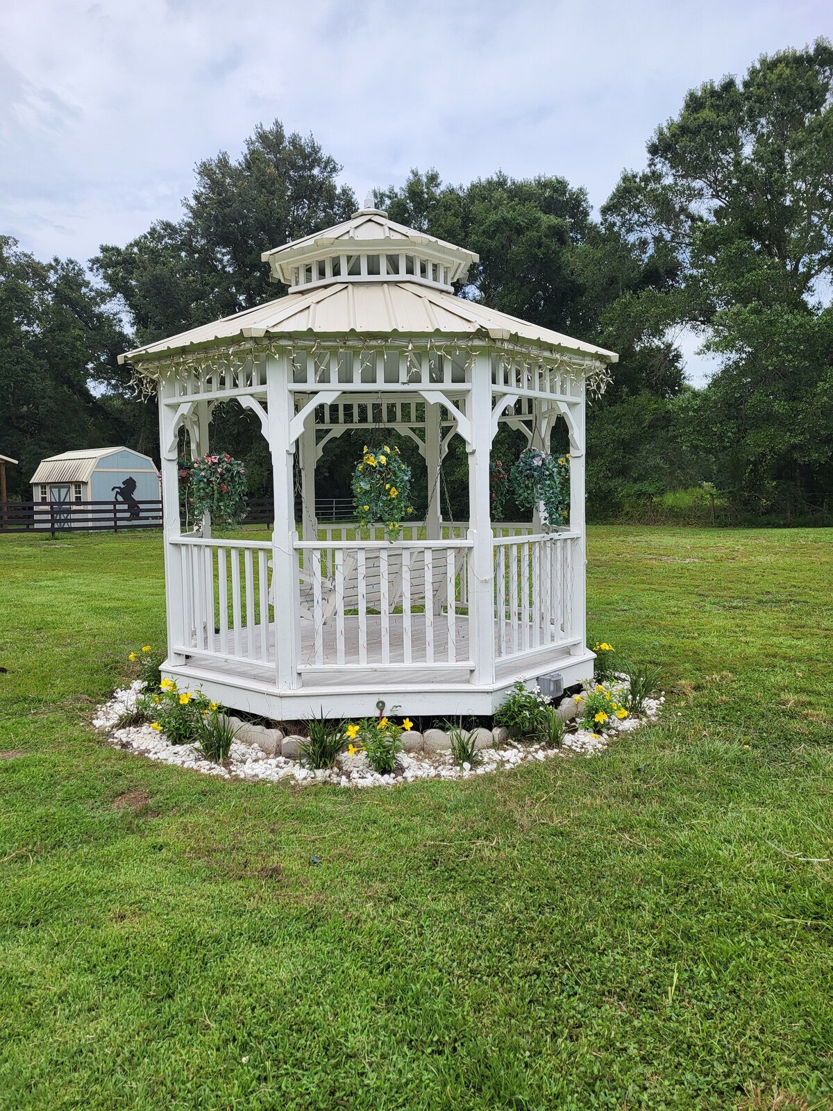 A white gazebo with a scalloped roof is situated on a grassy area surrounded by decorative stones and blooming flowers. The structure features open sides with vertical wooden slats, providing a spacious and airy feel. Lush greenery is visible in the background.