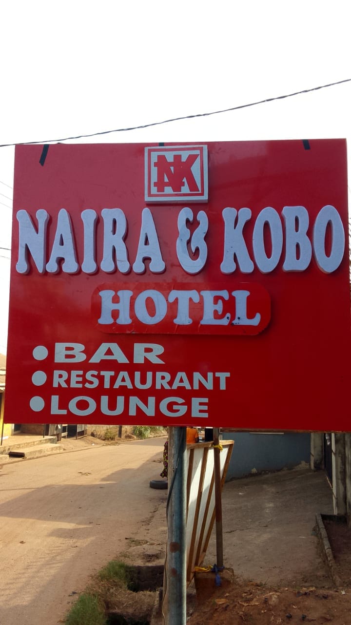 A vibrant red sign is displayed, featuring the name 'Naira & Kobo Hotel' prominently at the top. Below, the words 'Bar,' 'Restaurant,' and 'Lounge' are listed in white lettering, indicating the amenities available to guests. The sign is positioned outside in a relaxed urban setting.