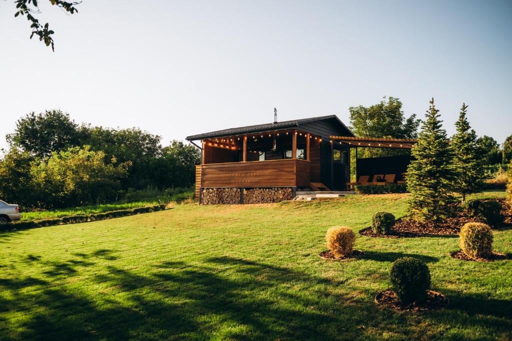 A single-story house with wooden exterior and a covered porch is surrounded by a well-maintained lawn. Trees and shrubs frame the property, while string lights add a subtle glow. A gravel path leads to the entrance, enhancing the outdoor space's inviting feel.