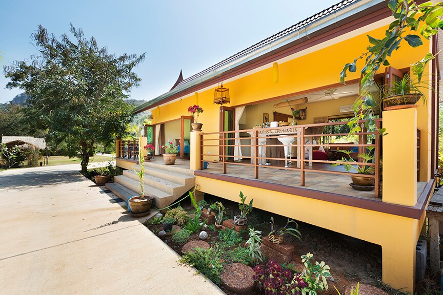 The exterior of a traditional Thai wooden house is showcased, featuring a vibrant yellow facade. A spacious terrace wraps around the front, adorned with potted plants and comfortable seating. Lush greenery surrounds the house, enhancing the tranquil garden setting.