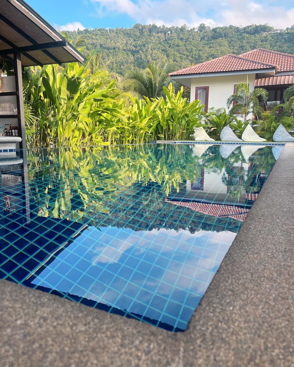The image features a reflective swimming pool surrounded by lush greenery and tropical plants. The serene water surface mirrors the vibrant blue sky above, with a modern structure visible in the background, showcasing a harmonious blend of nature and architecture.