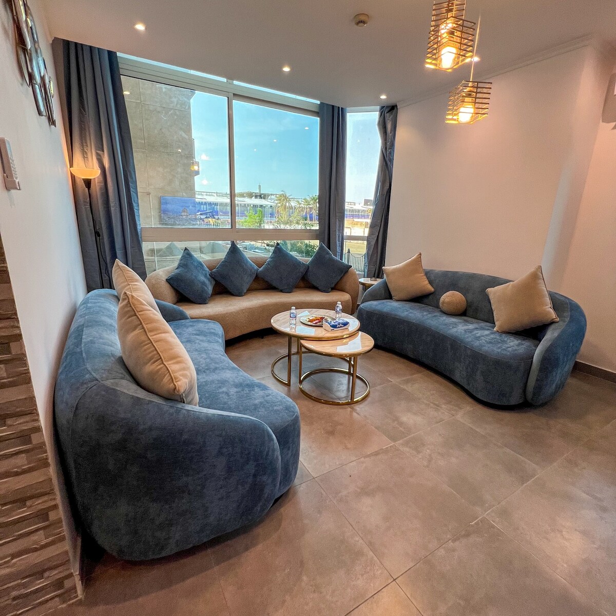 A modern living room is depicted, featuring a curved blue sofa adorned with various patterned cushions. A round coffee table sits in the center, displaying drinks. Large windows allow natural light to fill the space, offering views of the outside environment.