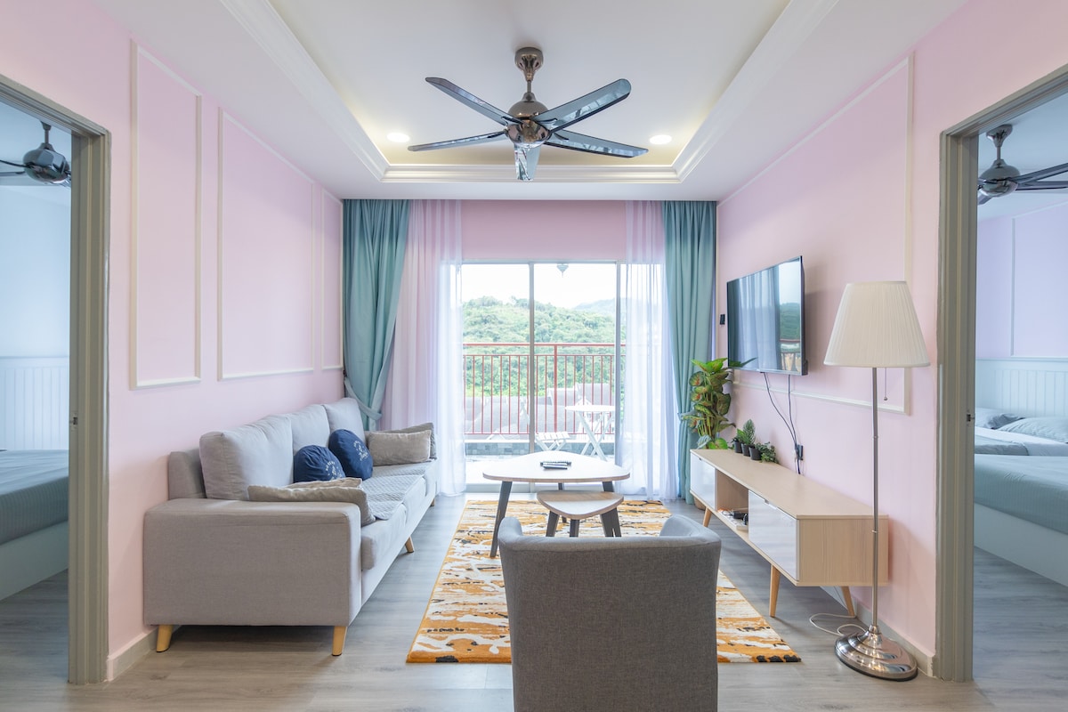 A spacious living area features soft pink walls and large windows that open to a balcony with scenic views. A comfortable gray sofa is positioned adjacent to a light wooden coffee table, with a relaxing rug beneath. A smart TV and a standing lamp add modern touches to the space.