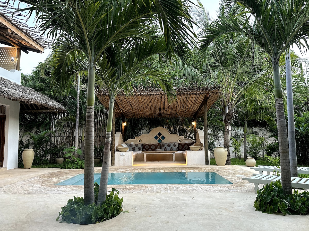 A tranquil outdoor area features a swimming pool surrounded by lush plants. A thatched gazebo with comfortable seating is positioned nearby, along with tall palm trees that provide shade and a serene ambiance.