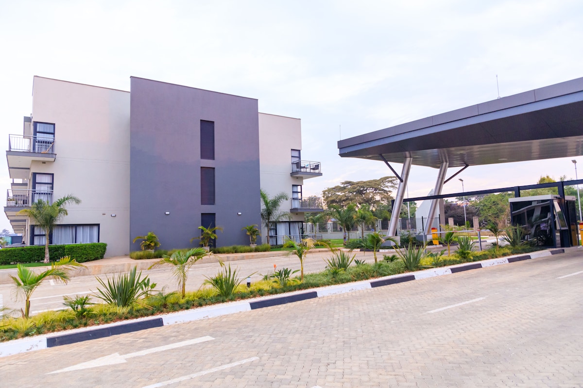 The modern apartment building is positioned alongside a neatly landscaped driveway. Tall palm trees and low shrubs enhance the exterior's appeal. A covered parking area can be seen, offering convenience for guests. The architecture features a blend of light and dark surfaces, creating a contemporary look.