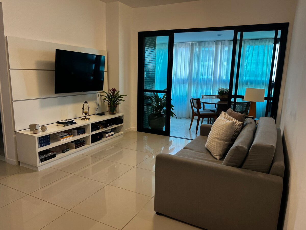 A modern living area features a sleek television mounted on a white wall unit. A comfortable gray sofa is positioned to face the screen, while a small dining space with a table and chairs is visible through large glass doors leading to a balcony.