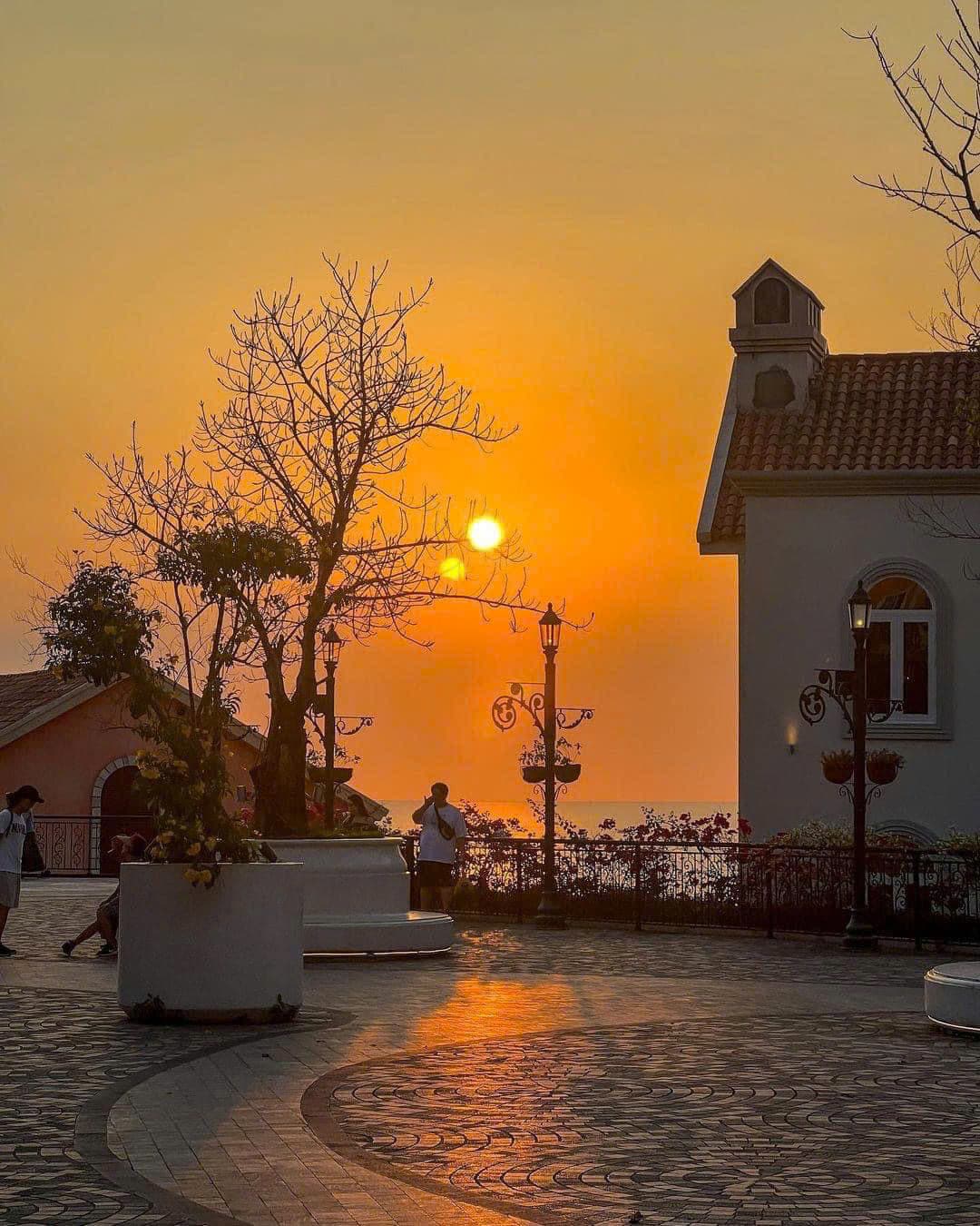 A tranquil scene showcases a sunset over the ocean, casting warm hues across the sky. Silhouettes of trees and a building frame the view, while pathways are lined with subtle lighting and decorative features, creating an inviting atmosphere for evening strolls.