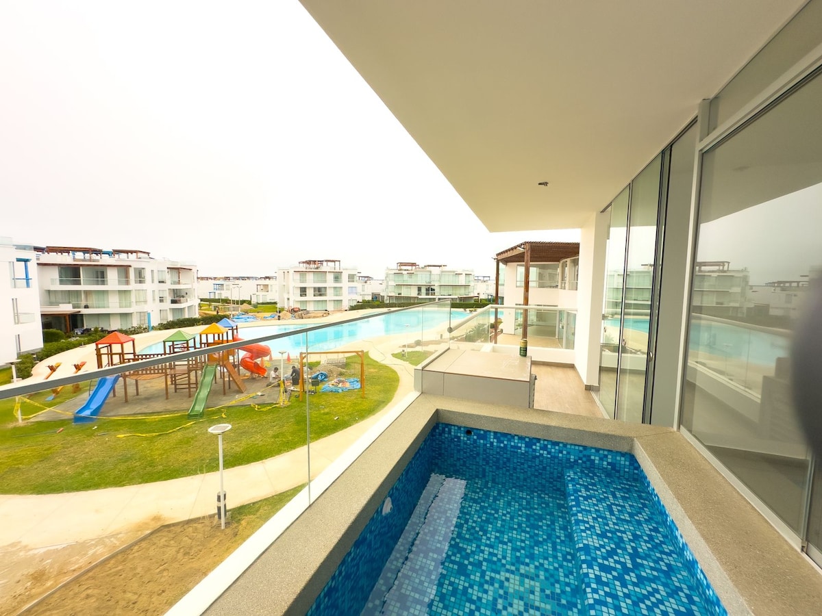 A view from the balcony shows a sparkling blue pool surrounded by landscaped green areas. A playground with slides and climbing structures can be seen nearby, providing recreational space. Expansive glass doors lead into the interior, emphasizing an abundant flow of natural light.