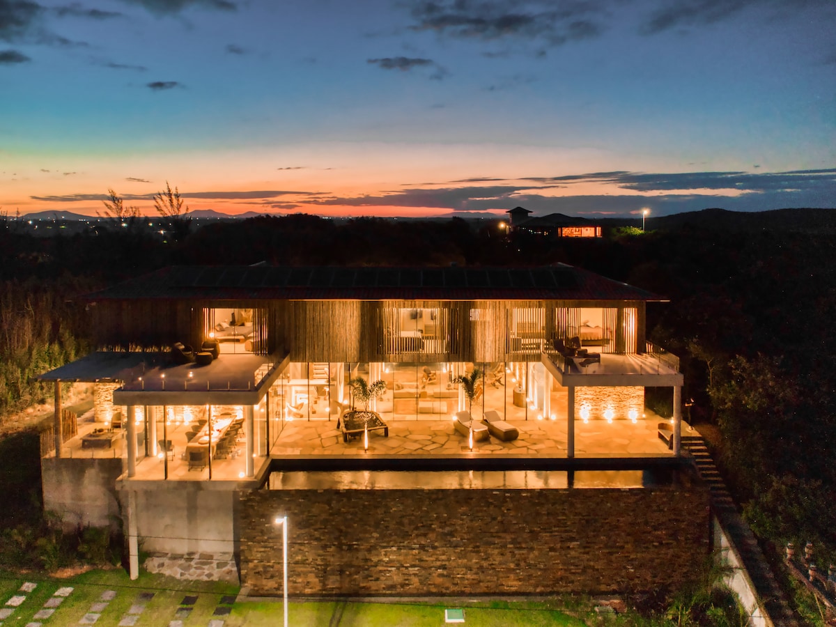 An aerial view of a modern private residence showcases large glass windows framed by natural elements. Warm lights create an inviting glow against the twilight sky, while the surrounding landscape features manicured green lawns and distant outlines of hills.