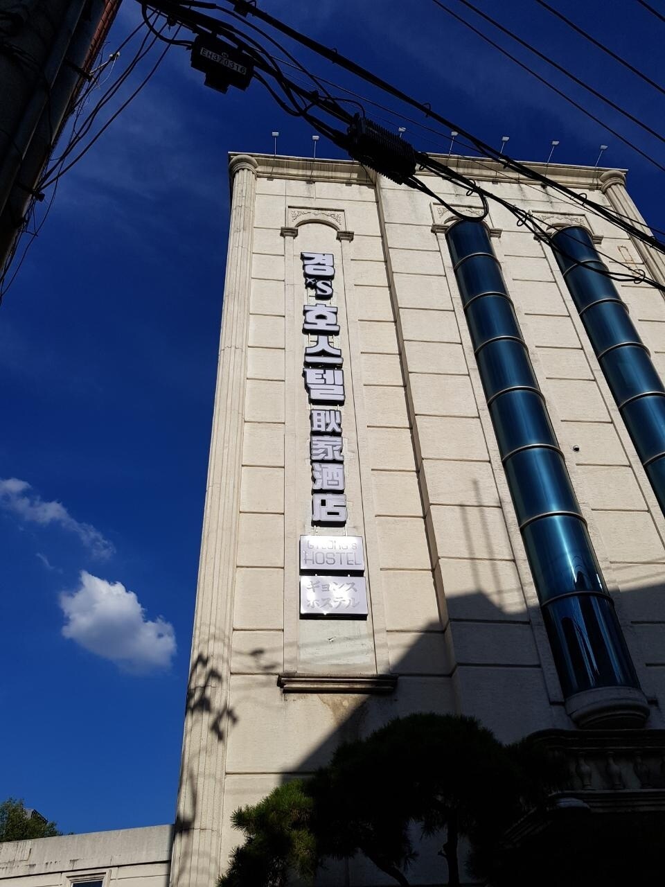 The exterior of a multi-story building is captured against a sunny blue sky. The facade features large glass windows and prominent signage indicating the establishment's name. A clear view of utility lines can be seen in the foreground, adding an urban context to the scene.