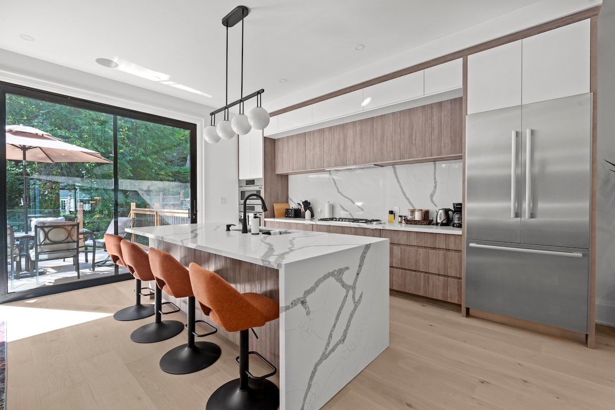 A modern kitchen features a large island with four orange stools and sleek cabinetry. Stainless steel appliances and a gas stove are visible. Floor-to-ceiling sliding glass doors provide access to an outdoor patio area, enhancing the spacious feel of the room.