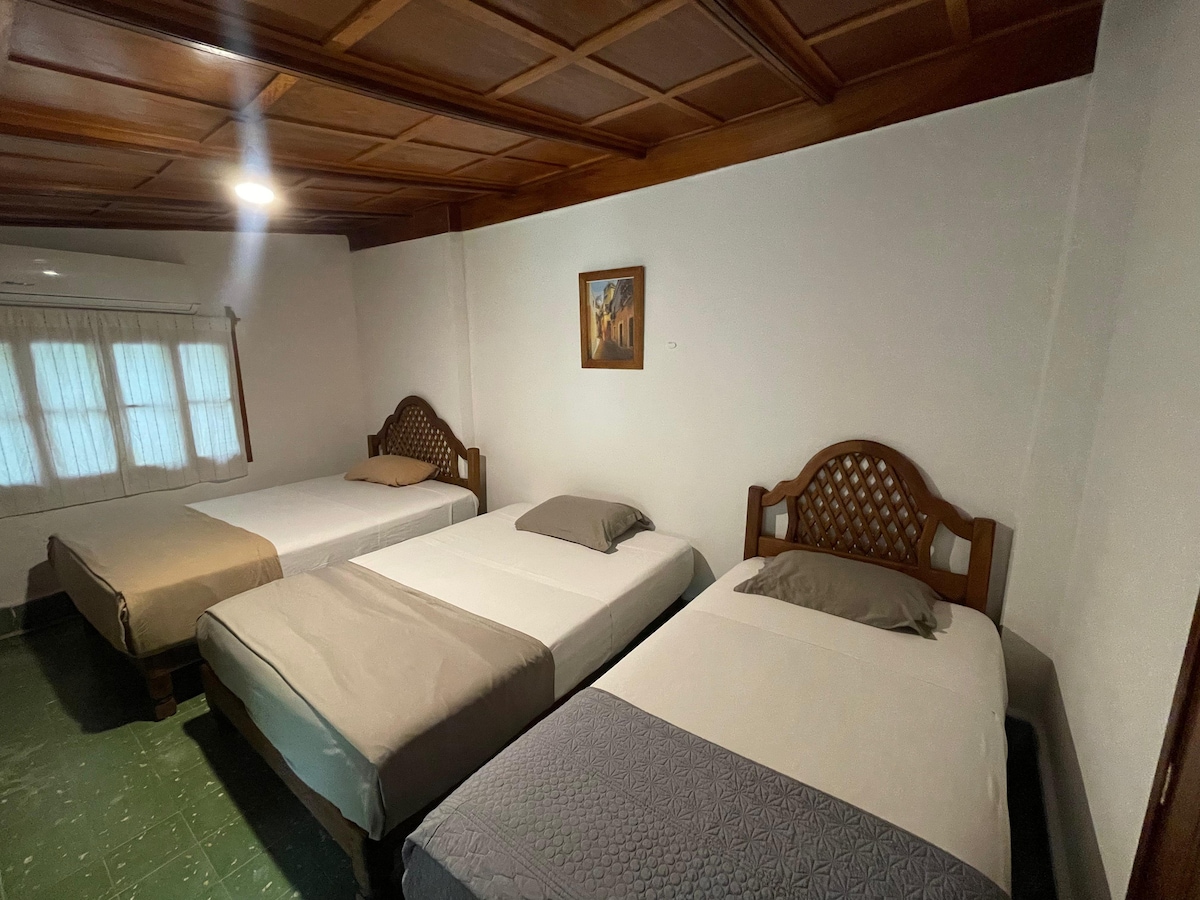A sleeping area is furnished with three individual beds, each featuring simple bedding in neutral tones. A piece of artwork hangs on the wall above one bed, while natural light enters through a window adorned with sheer curtains.