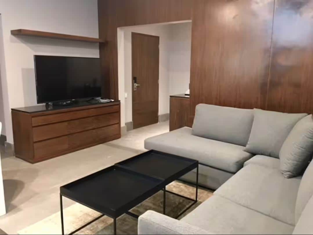 A modern living area features a light gray sectional sofa positioned next to a black coffee table. A large flat-screen TV is mounted on a wooden panel above a sleek storage unit. Natural light filters through a doorway leading to another space.