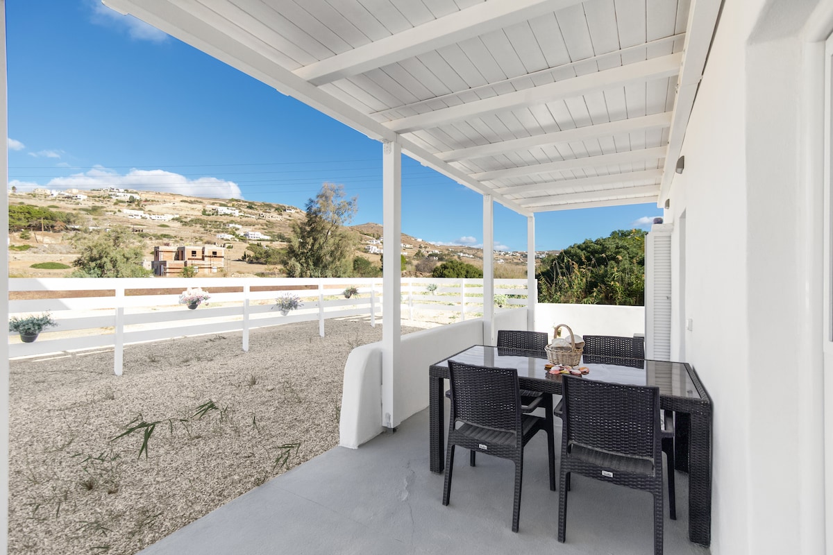 An outdoor terrace features a large dining table surrounded by black wicker chairs, set against a backdrop of rolling hills and clear blue skies. The area is shaded by a white wooden pergola, creating a comfortable space for relaxation and meals.