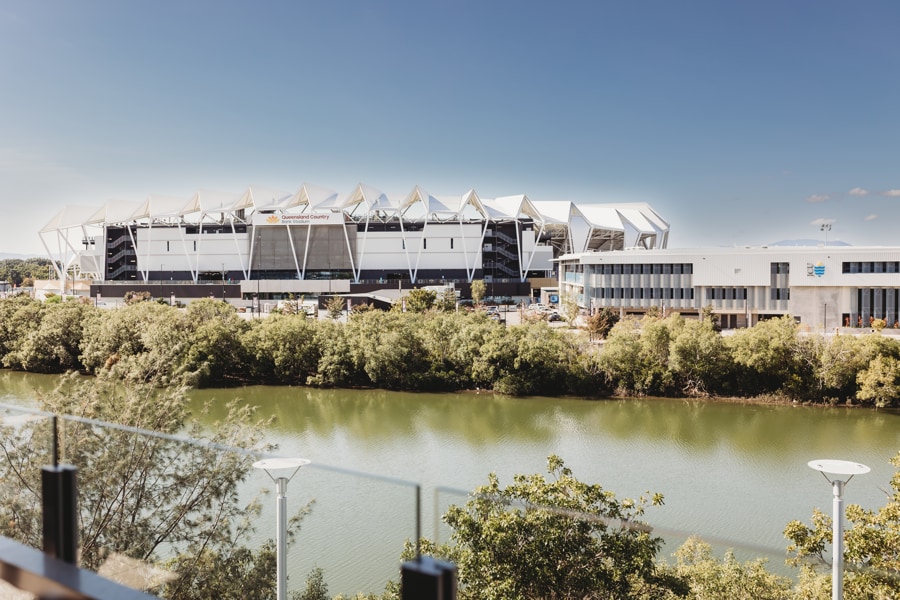 A scenic view of the QLD Country Bank Stadium, situated opposite the river. The stadium is complemented by the nearby modern building and lush greenery along the water's edge, providing a relaxing and open atmosphere.