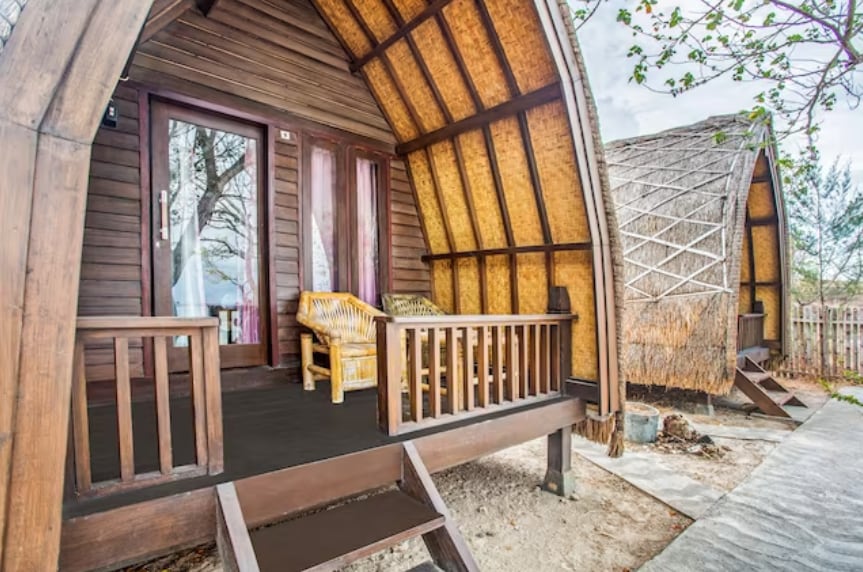 Wooden villas with distinctive curved roofs are set on a sandy path. A small porch features a seating area with natural wicker chairs, inviting relaxation. Lush greenery surrounds the entrance, enhancing the tropical ambiance.