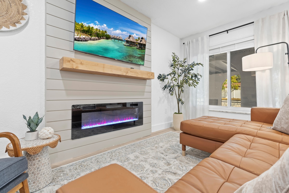 A modern living area features a tan leather sectional sofa positioned near a sleek electric fireplace. Above the fireplace, a television displays a serene beach scene. Light curtains frame a window, allowing natural light to illuminate the space, and a decorative plant adds a touch of greenery.