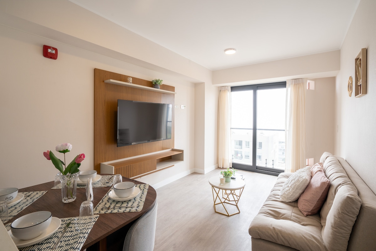 A bright living area is presented, featuring a comfortable sofa positioned near a wall-mounted television. A round coffee table sits in front of the sofa, complemented by elegant dishes and a vase with flowers on the dining table. Floor-to-ceiling windows allow natural light to fill the space.