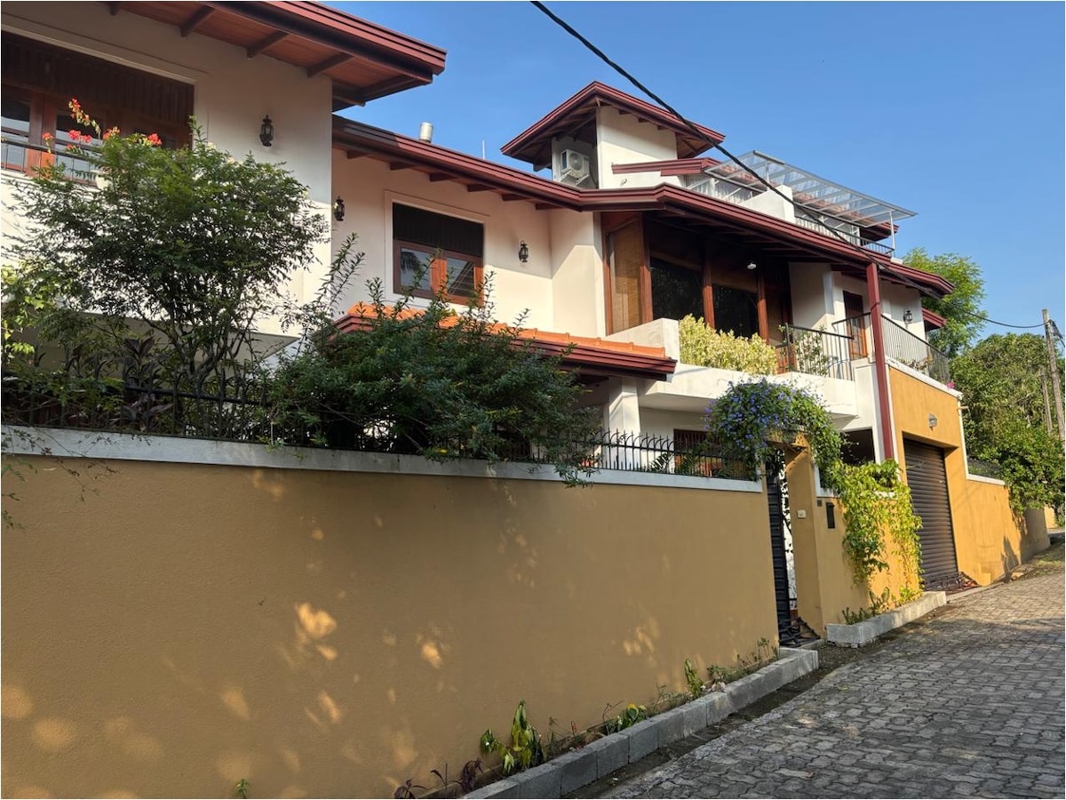 The exterior view of the apartment showcases a two-story structure set against a clear blue sky. A colorful garden with greenery enhances the entrance, while a pathway leads to the inviting front door. Architectural details provide a sense of modern elegance.