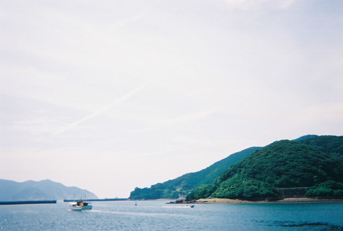 A serene coastal scene captures a boat navigating through calm waters, with rolling green hills in the background. The sky is mostly clear, and distant mountains create a gentle horizon, contributing to a peaceful atmosphere.