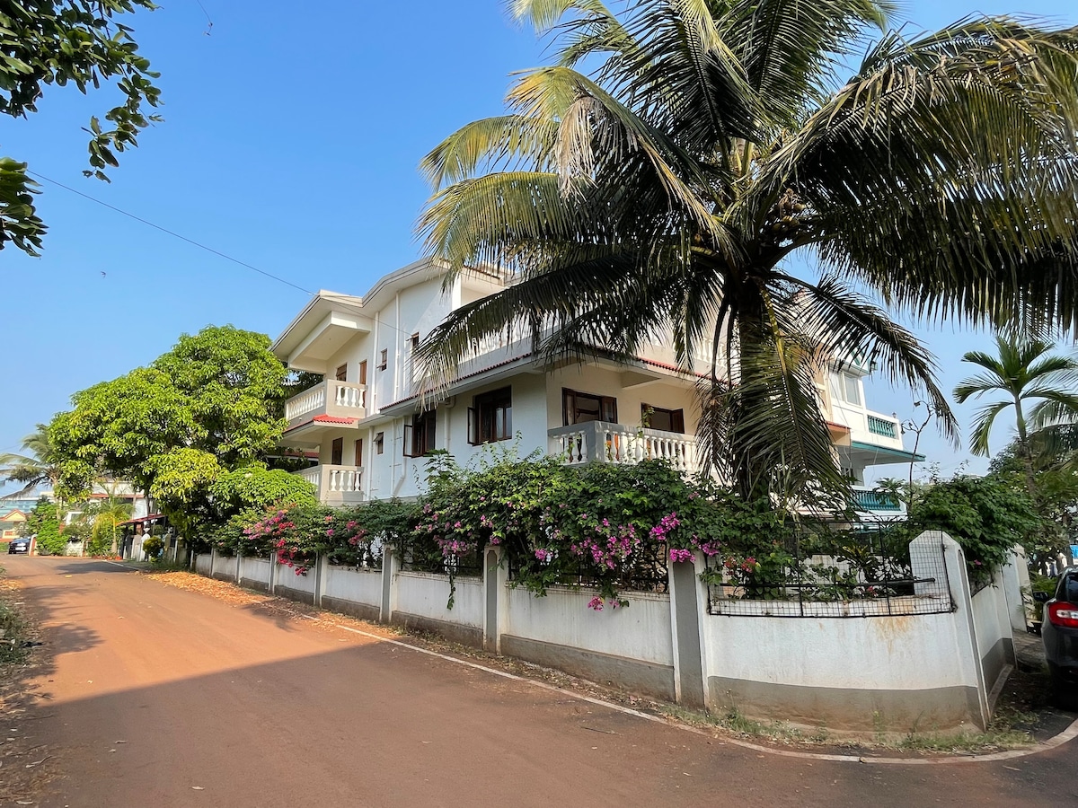 The exterior of the holiday let is set against a clear blue sky, surrounded by lush greenery. A well-maintained garden features flowering plants climbing the white walls, while palm trees provide shade. The building itself displays a modern architectural style with multiple balconies.
