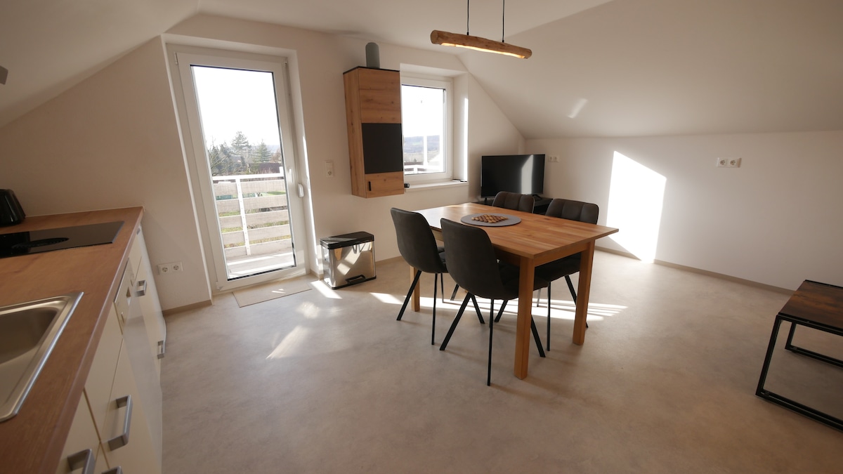 A bright living area incorporates a dining table surrounded by four chairs. Large windows provide natural light and access to a balcony. A modern kitchen with white cabinetry is visible in the background, alongside a sleek black armchair and a small table.