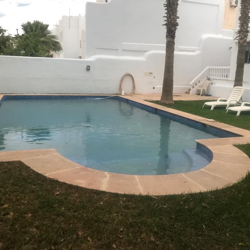 A private pool area is shown surrounded by well-kept grass and palm trees. The water reflects the sky, creating a serene atmosphere. A white lounge chair is positioned nearby, offering a spot for relaxation. The pool's rounded edges create a gentle, inviting shape.