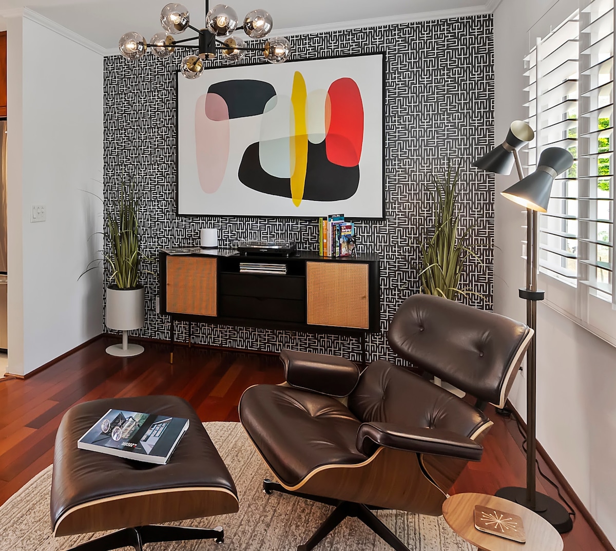 A modern living space showcases a stylish lounge chair with an ottoman, positioned near a sleek console table. A large abstract artwork adorns the wall, complemented by decorative plants and a floor lamp. Natural light filters through the window with wooden shutters.