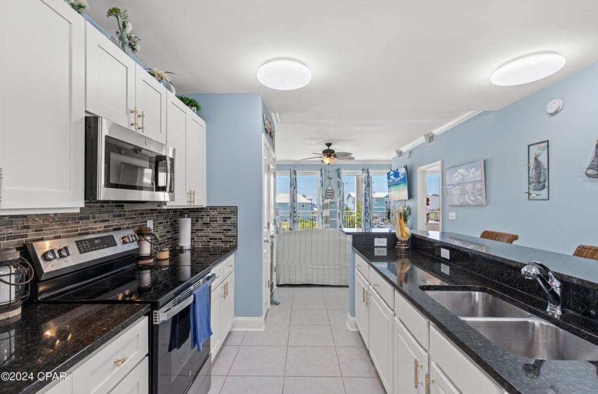 A bright kitchen features white cabinetry and a spacious black countertop. Modern stainless-steel appliances, including a microwave and oven, are visible. Natural light floods the area through a nearby window, while light blue walls create a calming backdrop.