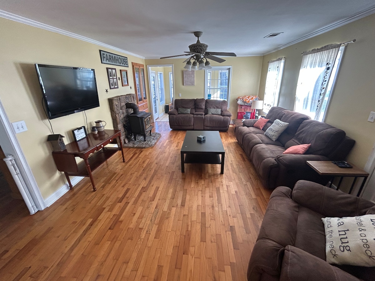 A spacious living area features two plush sofas and a matching loveseat arranged around a central coffee table. A television is mounted on the wall opposite, and a decorative fireplace adds warmth. Large windows allow natural light, while hardwood flooring offers a welcoming ambiance.