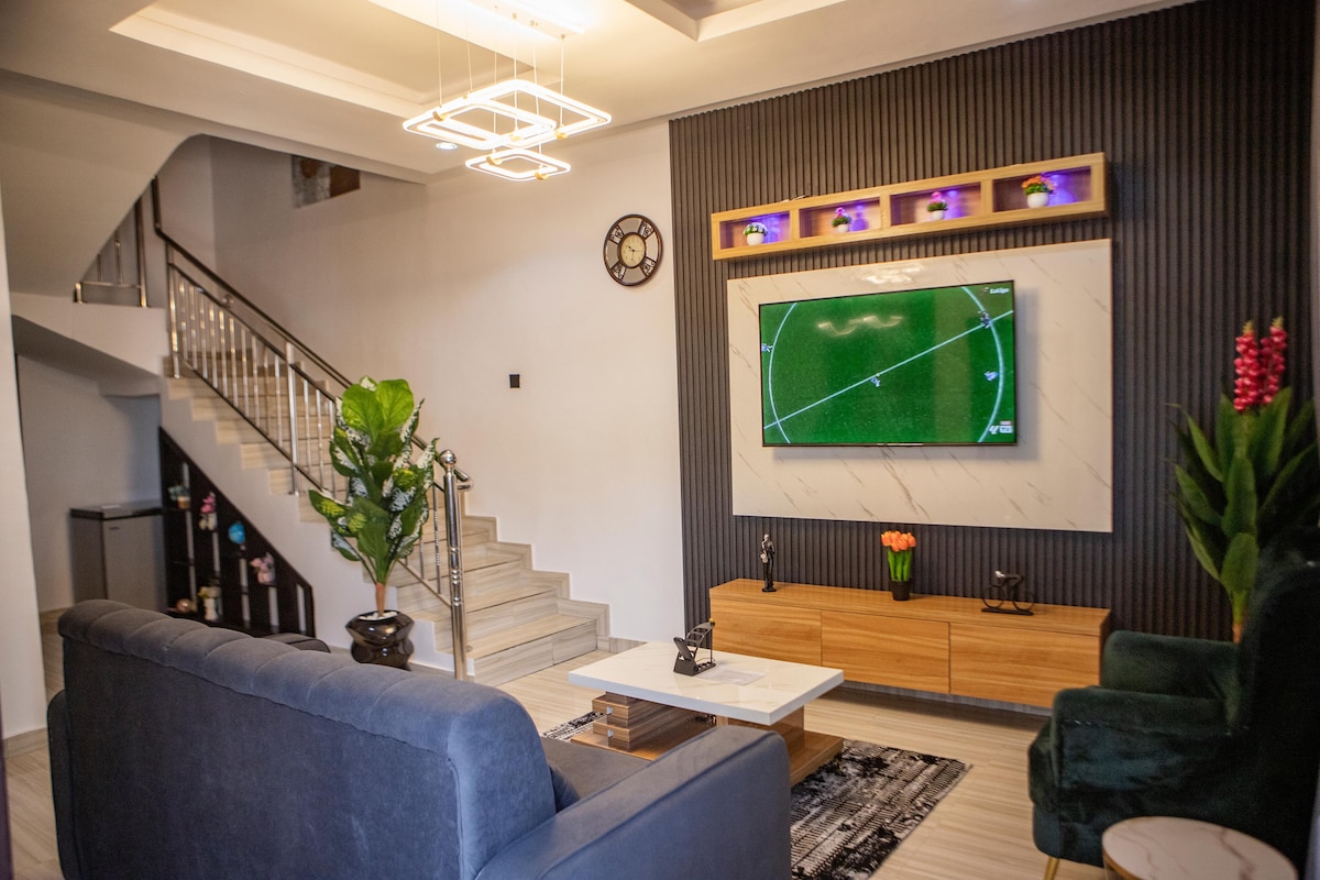 A modern living area is featured, showcasing a comfortable dark sofa and a sleek coffee table. A large wall-mounted TV displays a sports game. Decorative plants and soft lighting add a welcoming touch to the space. The staircase is visible in the background.
