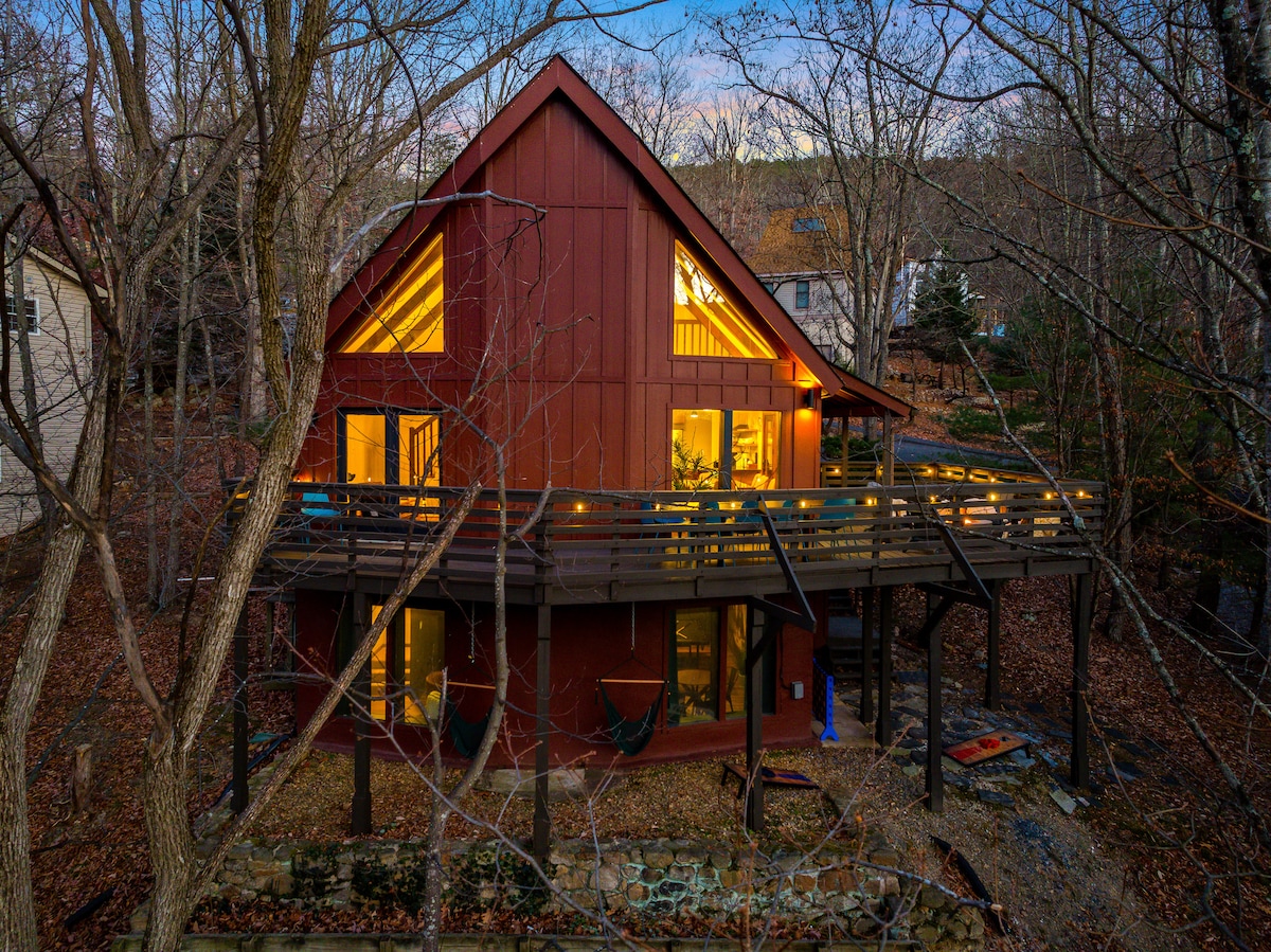 The exterior of the chalet is highlighted by a modern design with a sloped roof and large windows, allowing natural light inside. A spacious deck wraps around the building, featuring hammocks and surrounded by trees, creating a tranquil retreat in a natural setting.