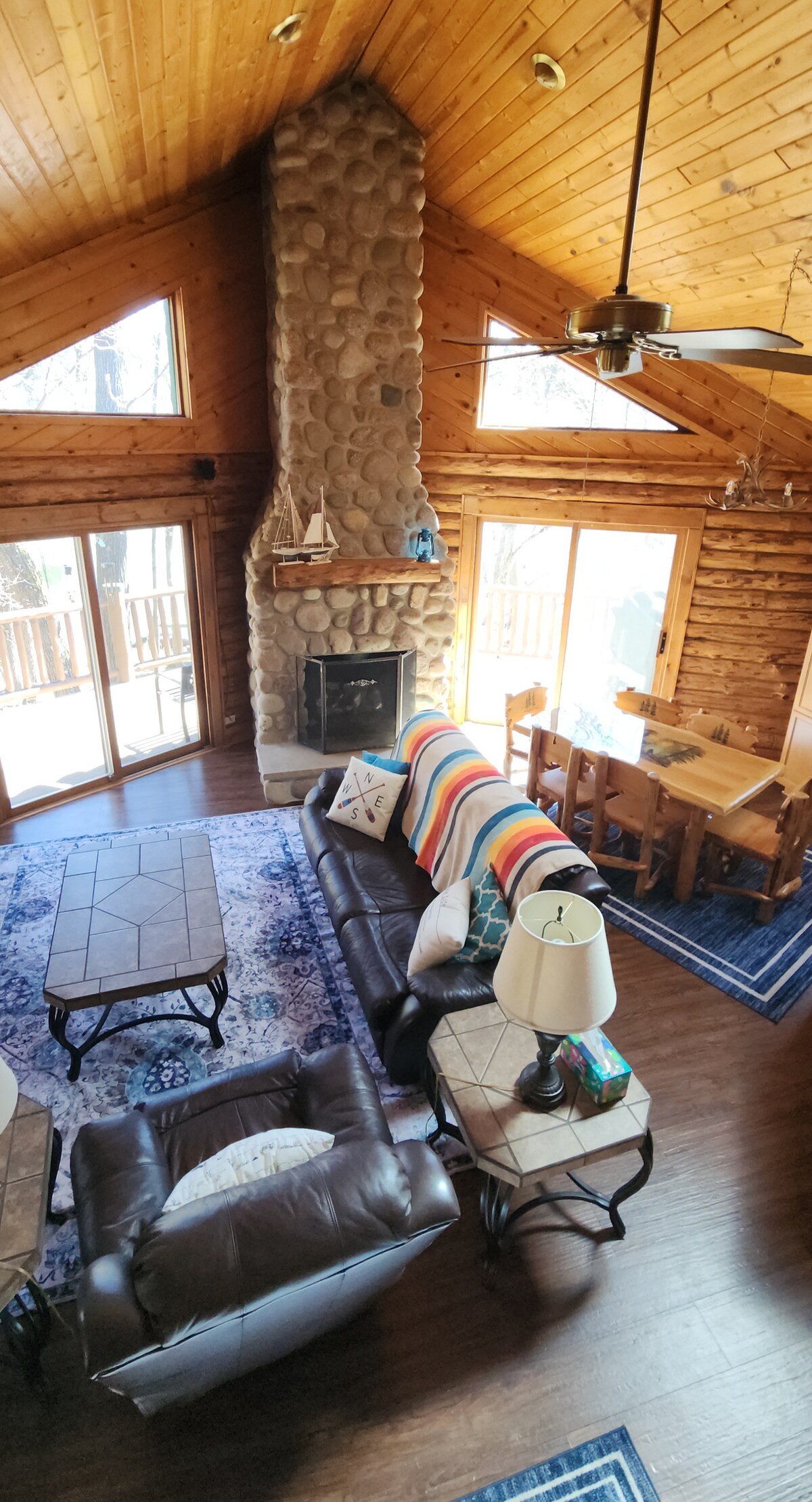 The cozy living area features a high wooden ceiling with a stone fireplace, enhancing the natural ambiance. Two coffee tables and a sofa are arranged for comfort, while a dining table sits nearby. Large glass doors provide access to an outdoor space with natural light.