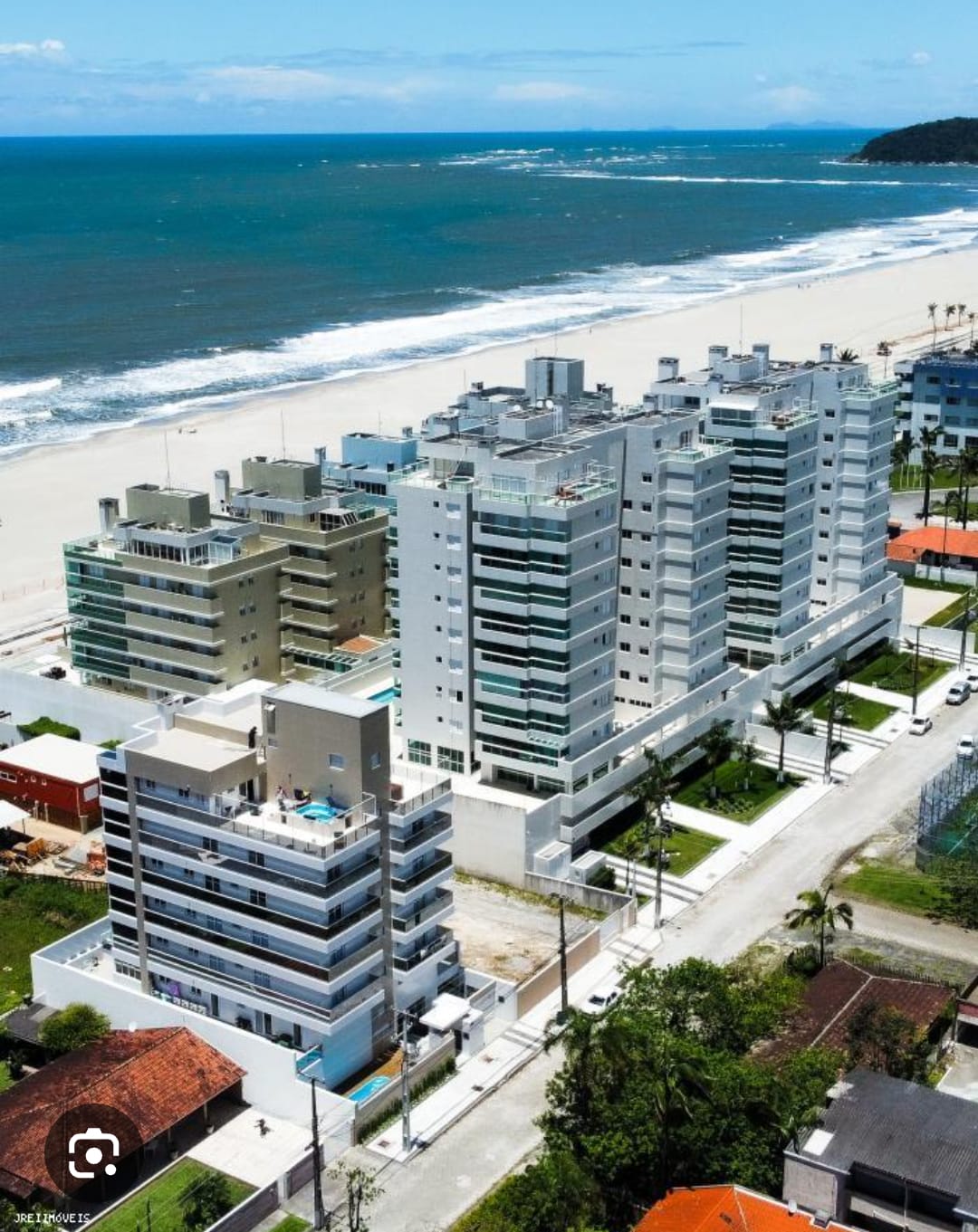 An aerial view captures multiple modern buildings along the coastline, set against the backdrop of the ocean. Sandy beach stretches out with gentle waves lapping at the shore, while palm trees line the streets, enhancing the coastal setting.