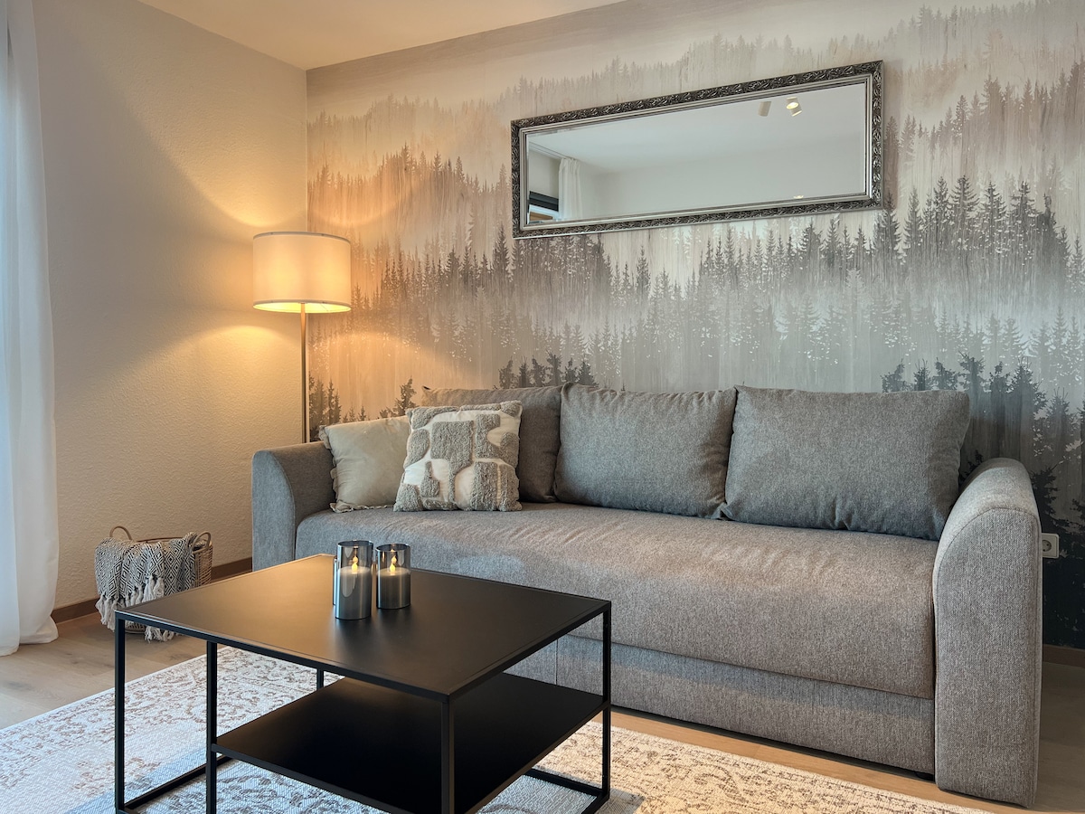 A spacious living area is highlighted by a gray sofa adorned with decorative pillows, positioned in front of a minimalistic black coffee table. A large mirror reflects the soft light from a nearby lamp, and a nature-themed mural creates a serene backdrop.
