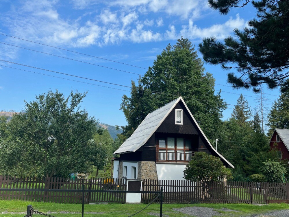 A charming, two-story cabin features a sloped roof and a blend of dark wood and stone accents. Surrounded by greenery, the property includes a wooden fence and is set against a backdrop of trees and distant hills under a clear blue sky.