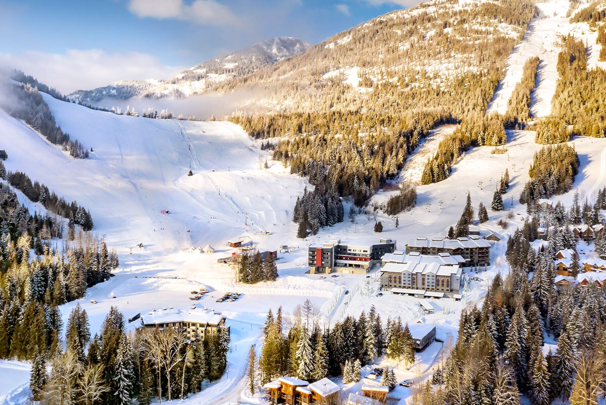 The image captures a panoramic view of a wintry landscape, showcasing snow-covered slopes and towering peaks. Resort buildings are nestled among trees blanketed in white, creating a serene setting. The bright sky complements the crisp, snowy terrain, enhancing the scene's tranquility.