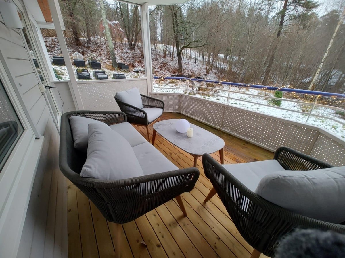 A covered outdoor seating area offers two woven armchairs and a round table. The wooden deck is surrounded by trees, with remnants of snow visible on the ground. Natural light enhances the serene setting, ideal for relaxation.