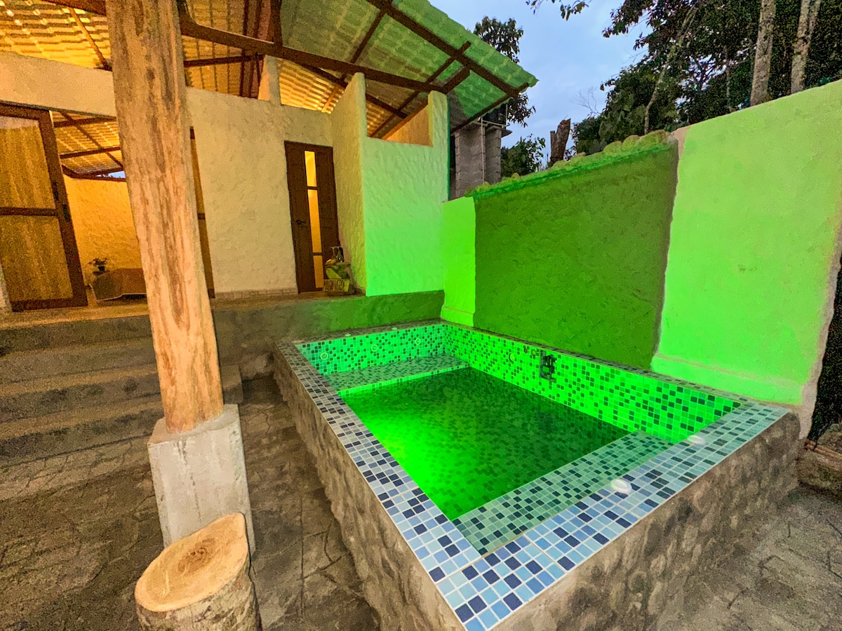 An outdoor jacuzzi is illuminated in soft green tones, surrounded by a tiled border. The stone flooring leads up to a rustic structure, combining natural materials with a modern touch. Its setting in a natural environment enhances the peaceful ambiance.