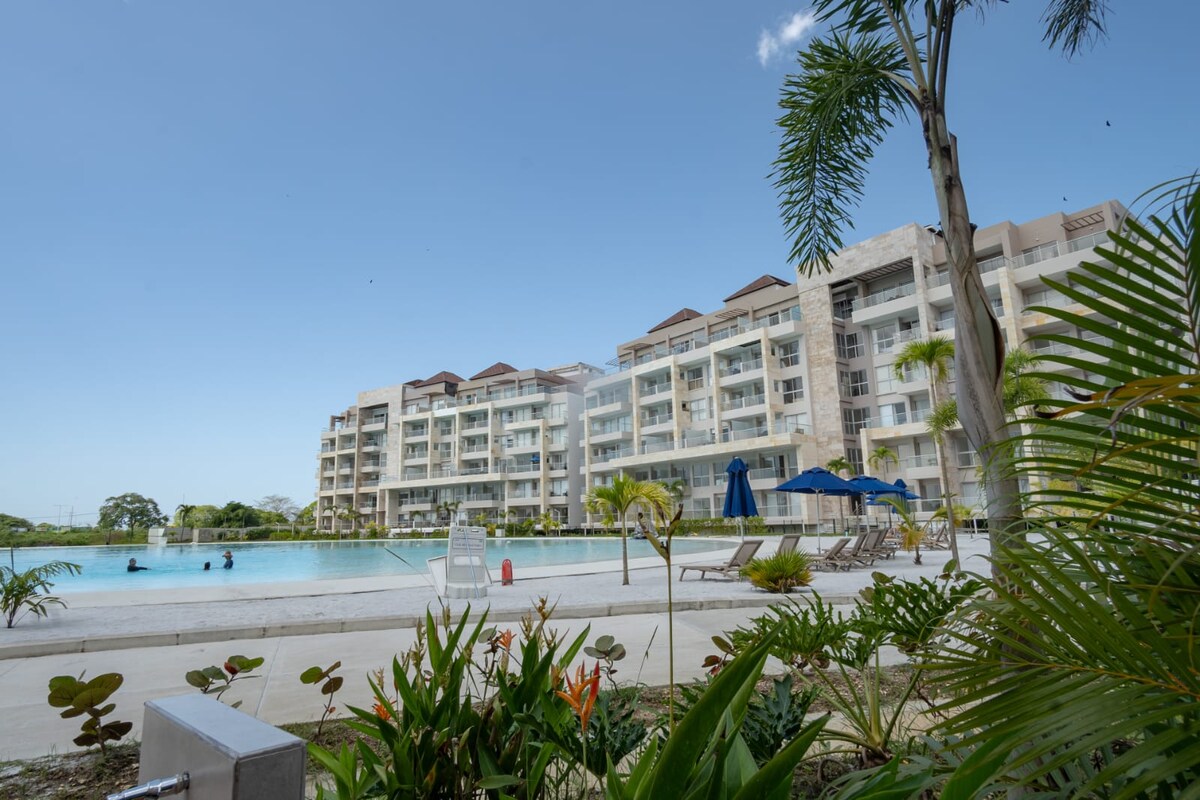 The image captures a modern condominium complex featuring multiple levels and spacious balconies. In the foreground, tropical plants and lounge chairs can be seen by a large, clear lagoon-style pool, while two umbrellas provide shade. Guests are visible enjoying the water.