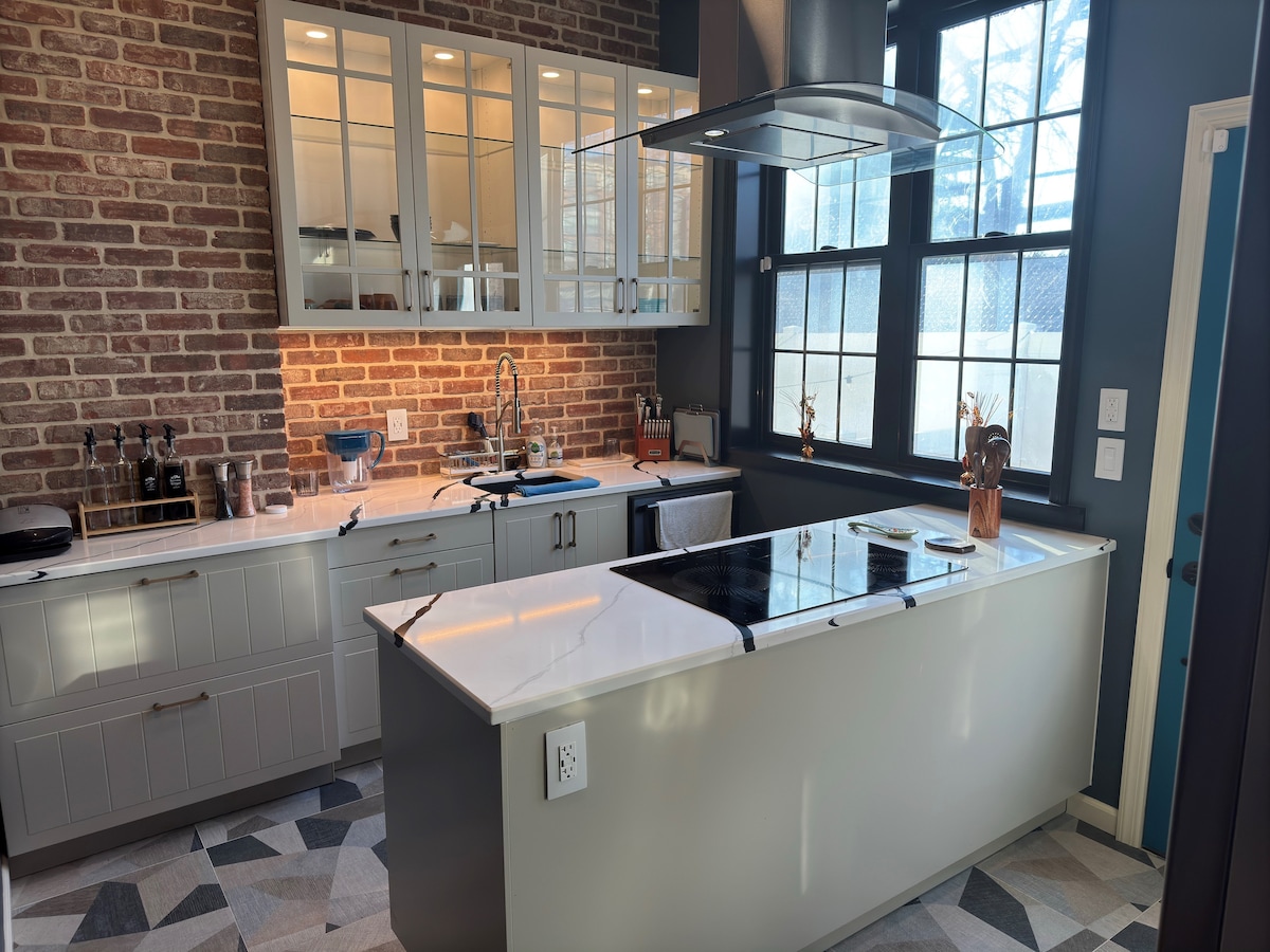 A modern kitchen features sleek cabinetry and a central island with an integrated cooktop. Large windows allow natural light to fill the space, accentuating the brick wall. The kitchen is equipped with various appliances and ample counter space, creating a functional cooking environment.