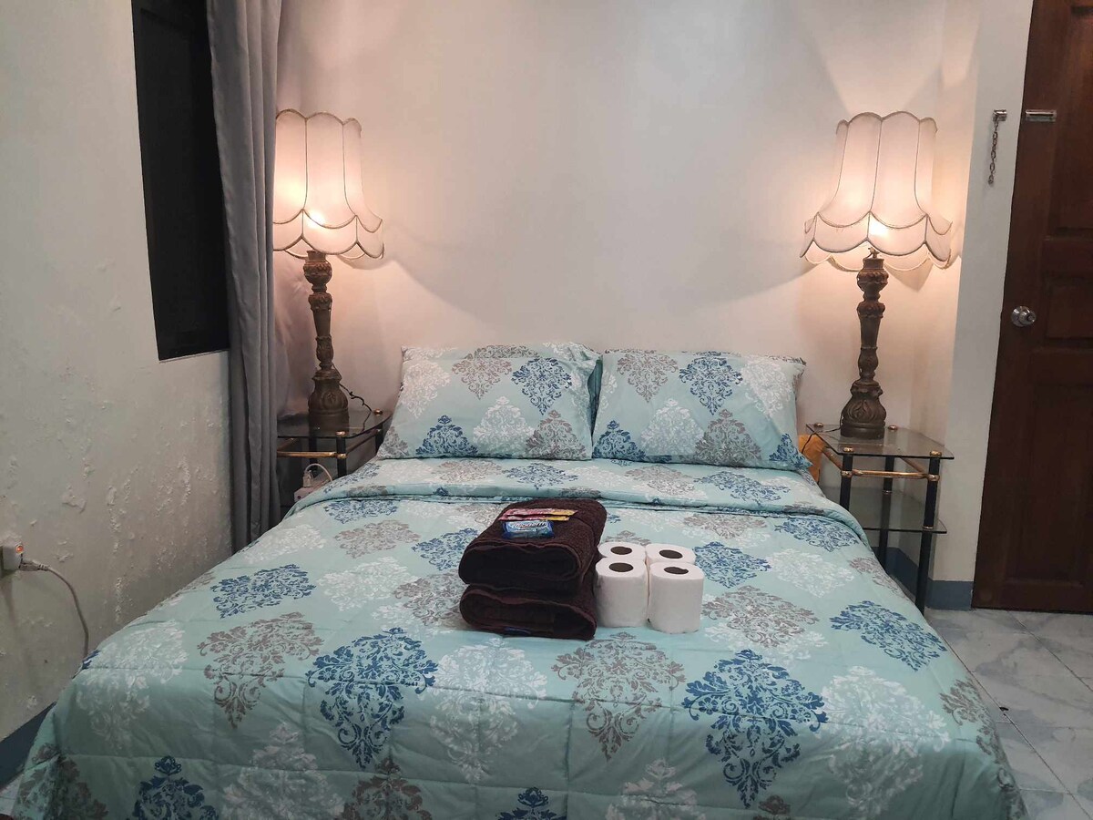A spacious bed is positioned in the center of the room, adorned with a turquoise and white patterned blanket. Matching bedside lamps cast a soft glow, and two decorative towels are neatly placed at the foot of the bed. Two rolls of toilet paper are arranged nearby.