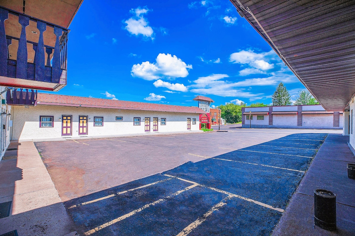 A spacious courtyard is displayed, featuring a well-maintained parking area. Surrounding the courtyard are single-story buildings, each with distinct doors. A clear blue sky adds brightness to the scene, while greenery is visible in the background.