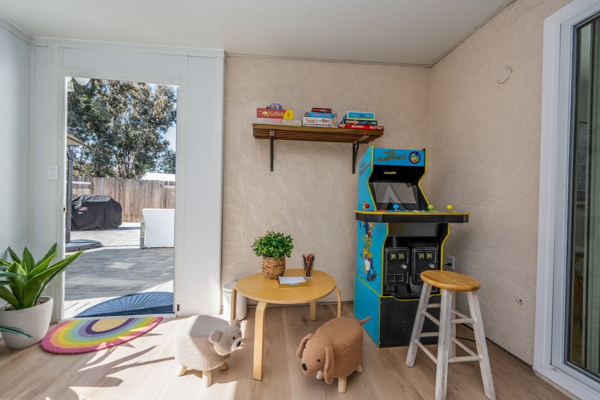 A bright game room features a colorful arcade machine and a small circular table surrounded by two plush animal stools. A shelf above holds various board games, and a potted plant adds a touch of greenery. Natural light fills the space from an adjoining door leading outside.
