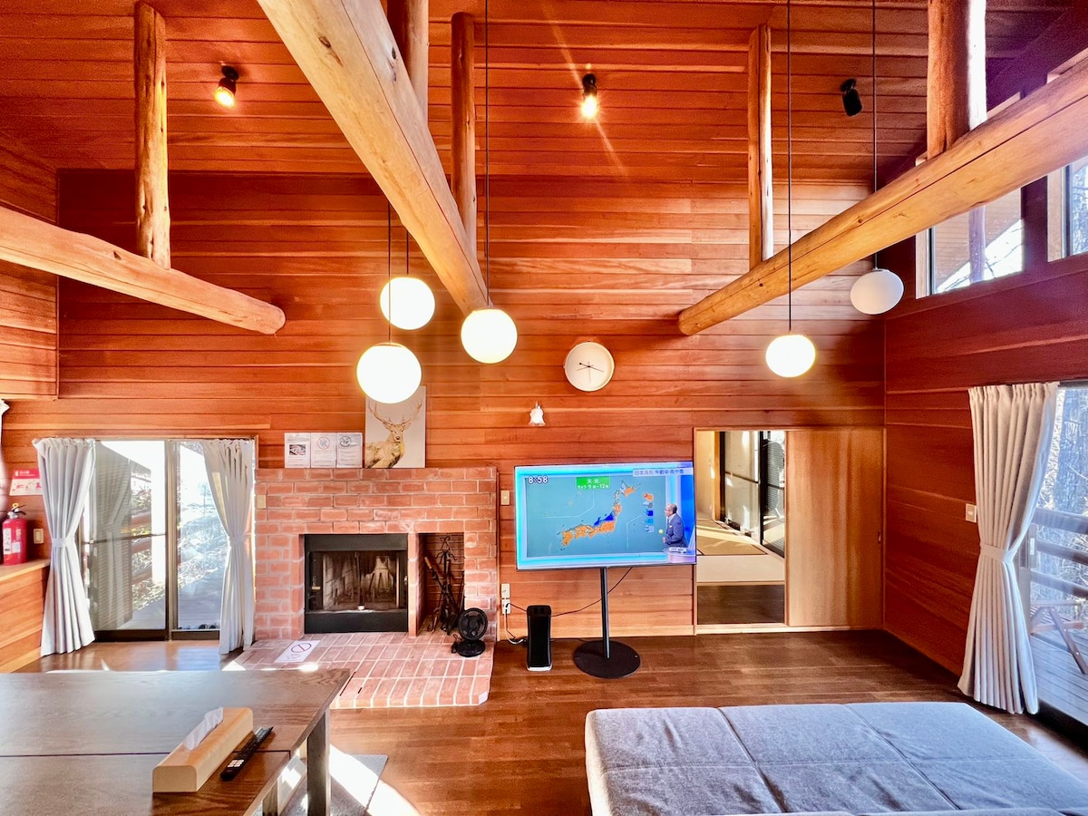 The living room features wooden walls and ceilings with exposed beams, creating a warm ambiance. A large flat-screen TV is mounted on a stand, facing a cozy seating area near a brick fireplace. Large windows allow natural light to fill the space.